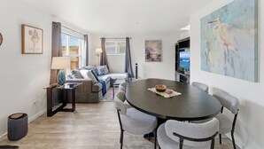 Een moderne woonkamer met een ronde tafel, grijze gestoffeerde stoelen, een bank en een schilderij aan de muur.