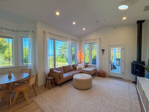 Ein geräumiger Wohnbereich mit einer Couch, Hocker und einem Esstisch mit Stühlen. Große Fenster mit Vorhängen, ein Kamin und eine Tür, die nach draußen führt.