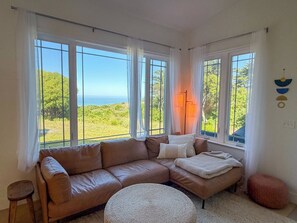 Ein Zimmer mit einer braunen Ledersofa, einem runden Hocker und Meerblick durch große Fenster.