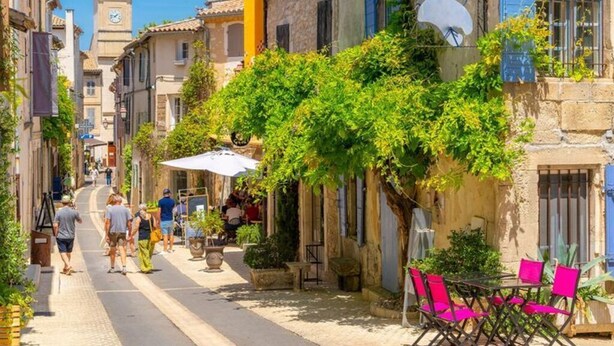 Uma rua estreita com edifícios em ambos os lados, mesas ao ar livre e uma torre do relógio ao longe.