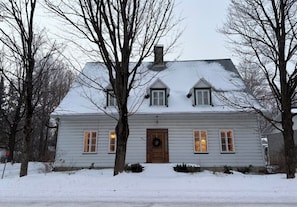Ein zweistöckiges Haus mit verschneitem Dach, einer Haustür mit Adventskranz und Fenstern mit Lichtern.