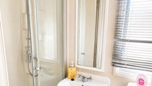 A bathroom with a glass shower enclosure, a white sink, a mirror, and a window with blinds. - 3 Bed Caravan - Sleeps 8 - Free Parking (Skegness)