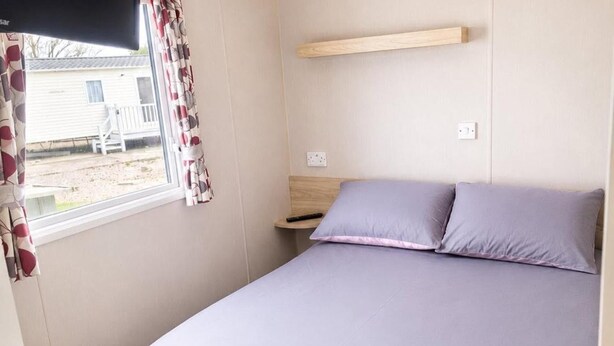 A single bed with a headboard, a bedside table, a power outlet, and a window with curtains. - 3 Bed Caravan - Sleeps 8 - Free Parking (Skegness)