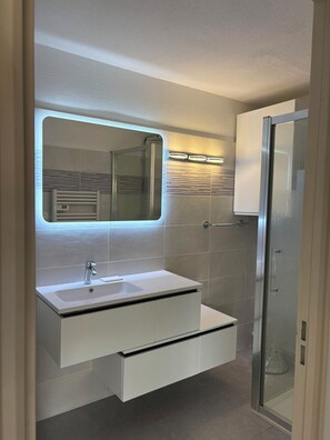 A modern bathroom with a large mirror, a built-in sink, a glass shower enclosure, and a towel rack. - Vrbo Property (Cavalaire-sur-Mer)