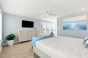 Ein Schlafzimmer mit Bett, Kommode, Fernseher, Deckenventilator, Fenster mit Jalousien und einem großen Gemälde.