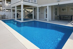 A rectangular swimming pool with a white deck surrounding it, a covered seating area with chairs, and a building with a balcony in the background. - BRAND NEW Luxury Semi-Oceanfront in Kill Devil Hills Seaduction (Kill Devil Hills)
