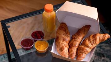 Una mesa de vidrio con una caja de cruasanes, un botellón de jugo de naranja y recipientes pequeños de mermelada y miel.