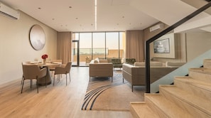 A modern living room with a dining area, a staircase, and a large window offering a city view. - Lafeef Apartments Al-Namuzji (Madinah)