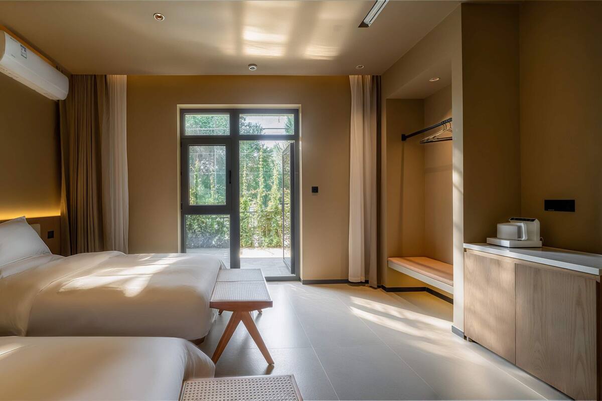 A hotel room with two beds, a desk, a coffee machine, and a view of greenery through the window.