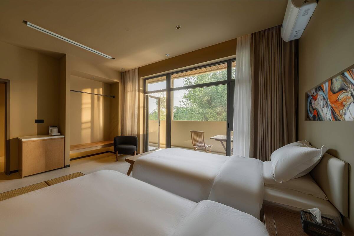A hotel room with two beds, a desk, a chair, a shower, and a large window with curtains.