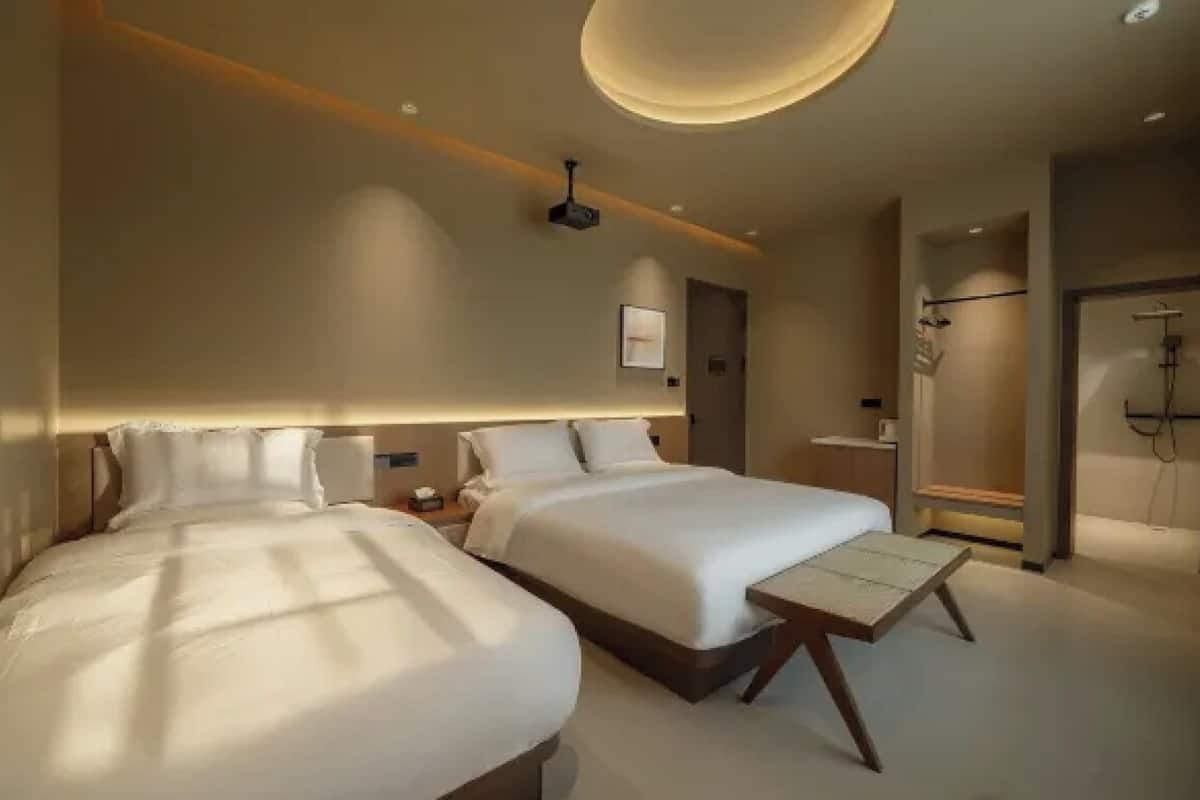 A hotel room with two beds, a small table, a shower area, and a ceiling light fixture.