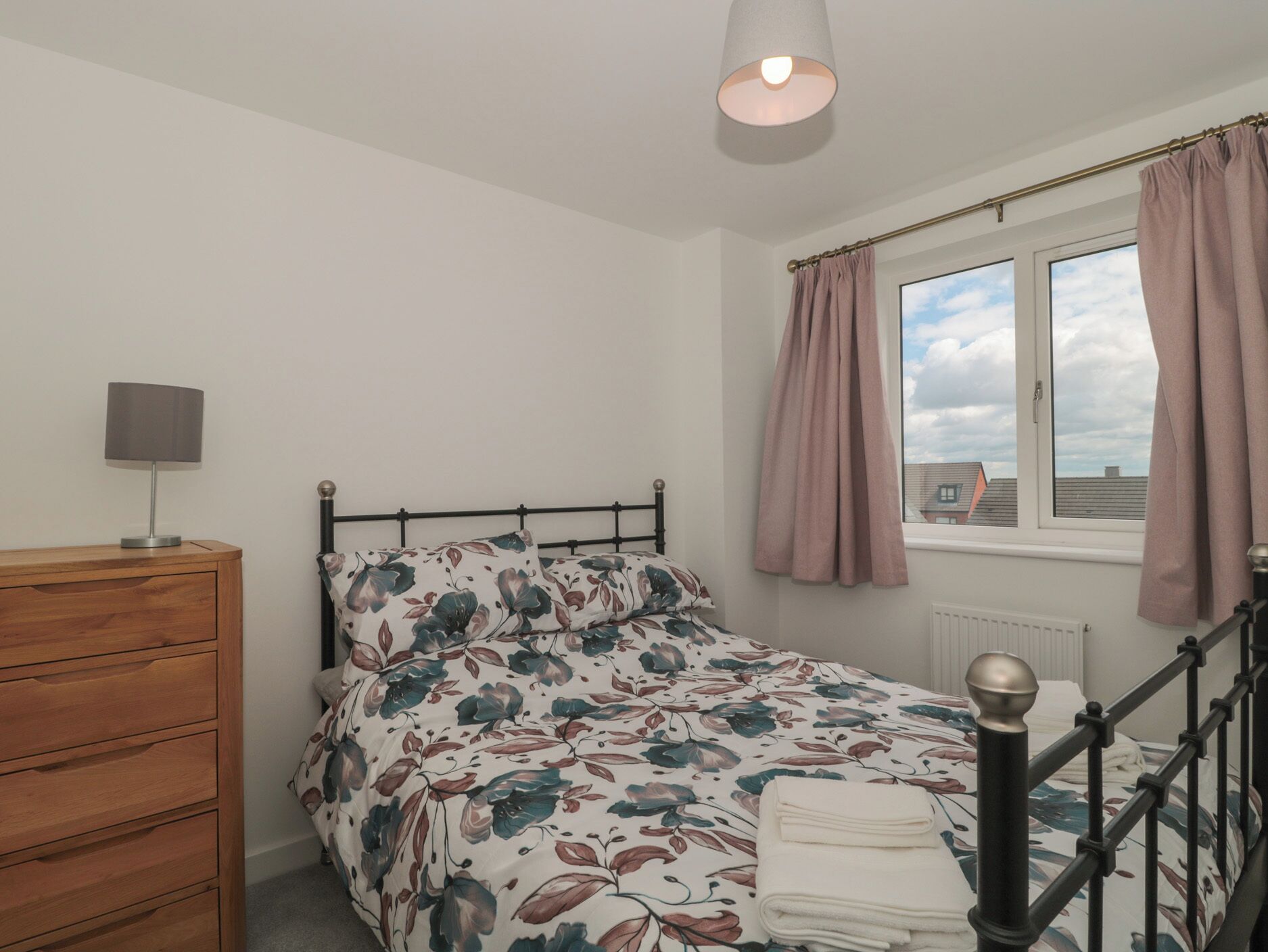 A bedroom with a bed, a wooden dresser, a lamp, a window with curtains, and a view of the sky.