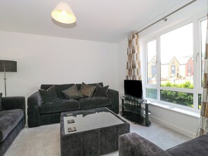 A modern living room with a sofa set, a coffee table, a TV, and a large window with a view of houses. - Beacon View (Malvern)