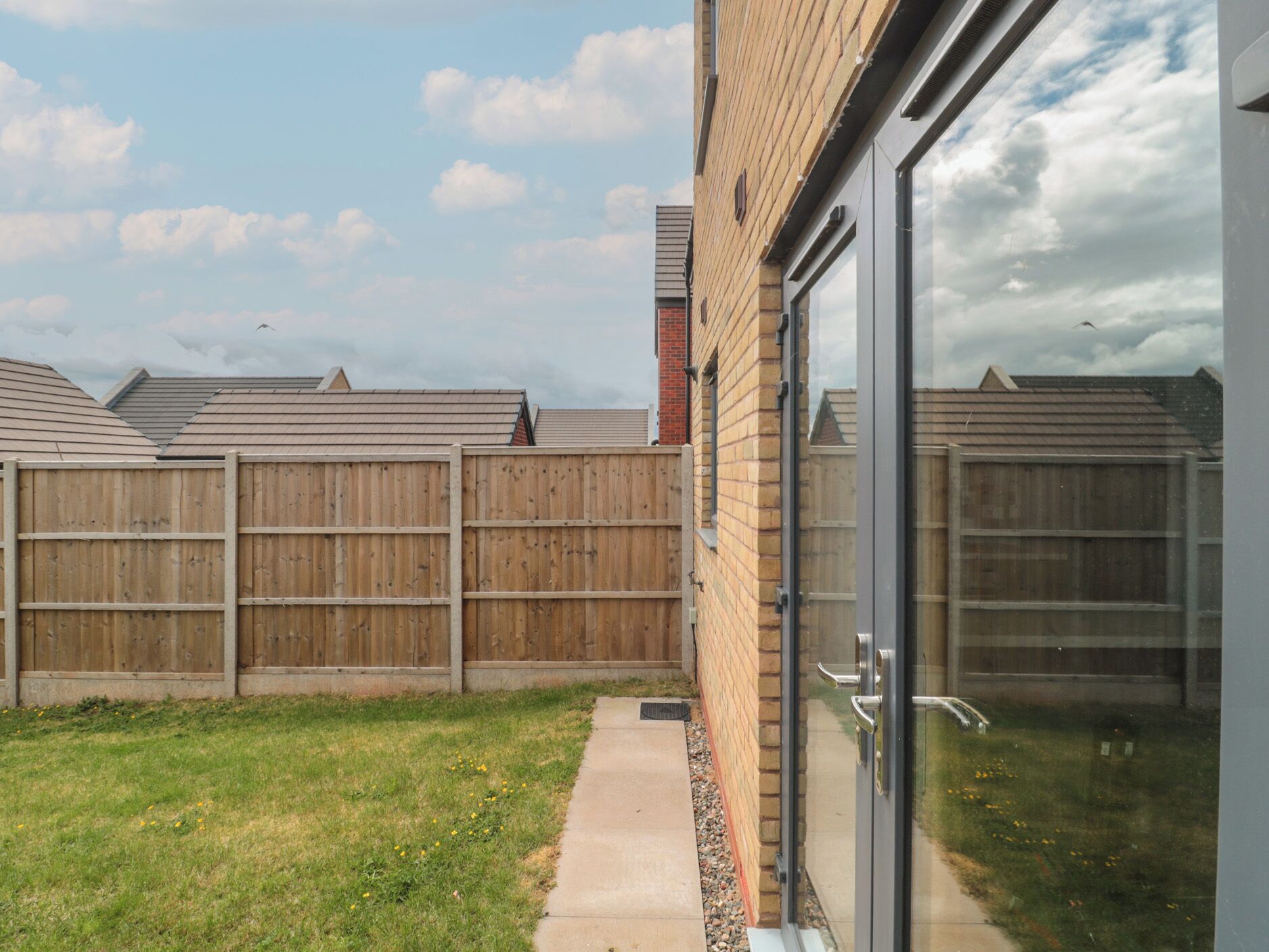A backyard with a wooden fence, a concrete pathway, and a brick building with a glass door.