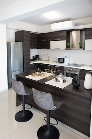 A modern kitchen with dark wood cabinets, a stainless steel refrigerator, a microwave, and a gas stove. There are two bar stools with a geometric-patterned placemat on the island.
