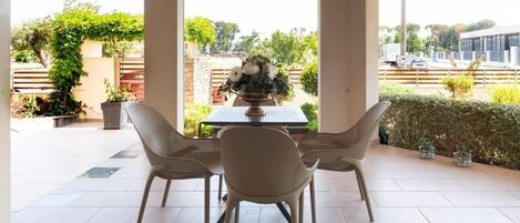 A covered patio with a table and chairs, a view of greenery and a building in the background.
