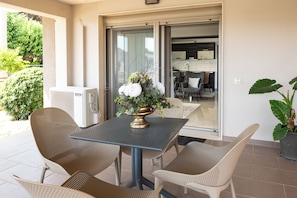 Um pátio externo com mesa e cadeiras, um vaso de planta e vista do interior por uma porta de vidro.