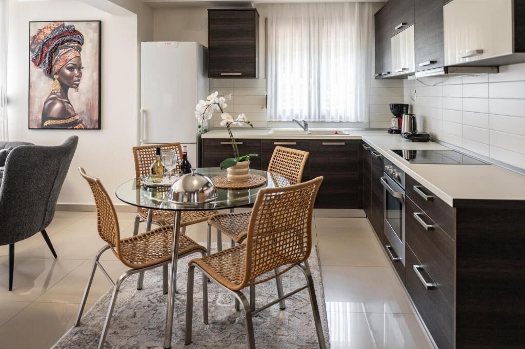 A modern kitchen with dark cabinetry, a glass dining table, wicker chairs, and a white refrigerator.