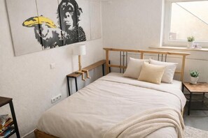 Ein Schlafzimmer mit Bett, Nachttisch, Lampe, Fenster und einer Wand mit einem Poster.