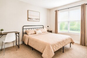 Ein Schlafzimmer mit Bett, Schreibtisch, Stuhl, Nachttisch, Lampe, einem Fenster mit Vorhängen und einem an der Wand hängenden Bild.