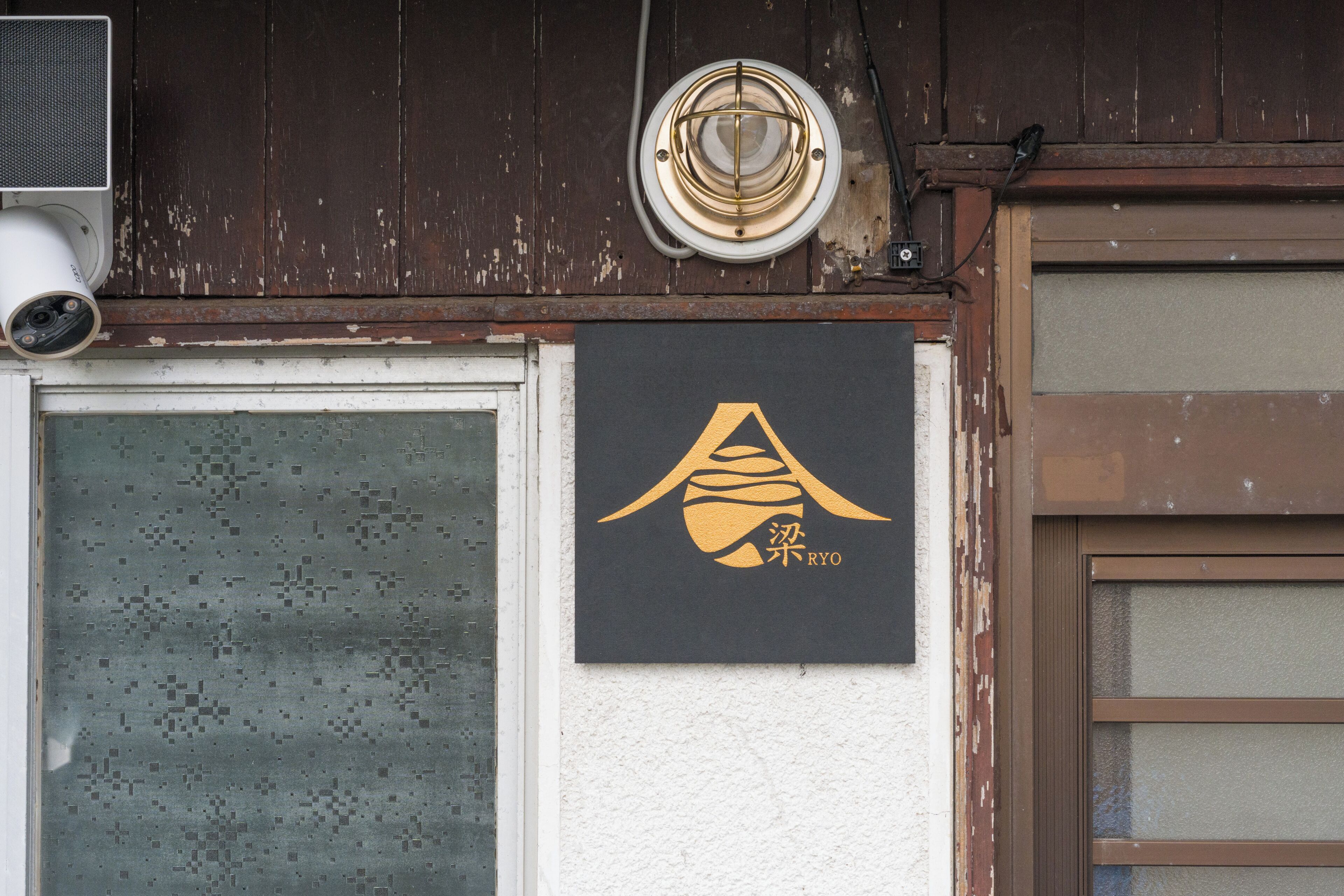 A building facade with a weathered brown wooden exterior, a security camera, and a black sign with gold lettering.