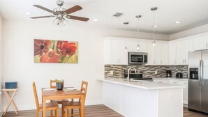 A modern kitchen with white cabinets, a stainless steel refrigerator, a microwave, and a coffee maker. There is a dining table with chairs and a ceiling fan. - Reed Unit B - Quiet Solo Room w/Shared Bath in Co-Living Home (Houston)