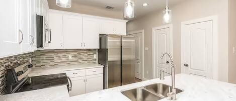 A modern kitchen with white cabinets, a stainless steel refrigerator, a marble countertop, and pendant lighting.