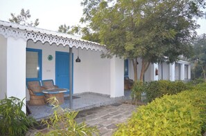 A building with a white exterior and blue doors, a small outdoor seating area with wicker furniture, and a tree in the background. - Farm Aravalia (Faridabad)