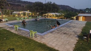 A swimming pool with surrounding chairs and a covered area, set against a hilly backdrop. - Farm Aravalia (Faridabad)
