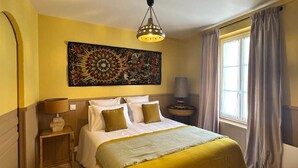Quarto de hotel com cama, mesa de cabeceira, luminária, janela com cortinas e um objeto decorativo na parede.