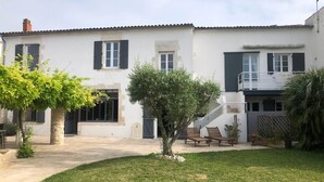 Uma casa de dois andares com fachada branca, persianas pretas e um pequeno jardim com uma árvore e um banco.
