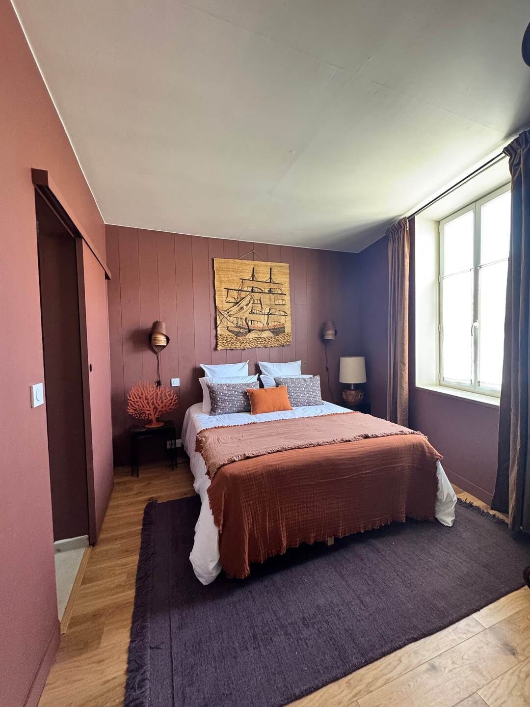 Un dormitorio con cama, una obra de arte en la pared, una mesita de noche, una lámpara y una ventana.