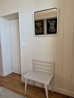 Eine weiße Bank mit querverlaufenden Leisten, vor einer weißen Tür und einer Wand, auf der sich ein kleines Spiegelbild zweier gerahmter Kunstwerke zeigt.