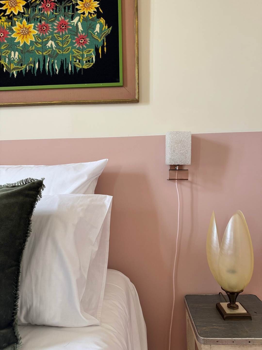 Een bed met witte en donkergroene kussens, een wandlamp en een bloemig kunstwerk in een lijst erboven.