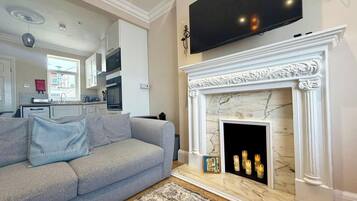 Ein Wohnzimmer mit grauem Sofa, Kamin inklusive Kerzen, einem darüber montierten Fernseher und einer Küche im Hintergrund.