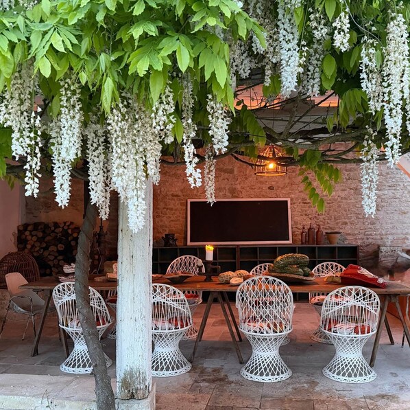Un espace de restauration extérieur rustique, avec des chaises en osier blanc, une table en bois et des fleurs blanches suspendues.