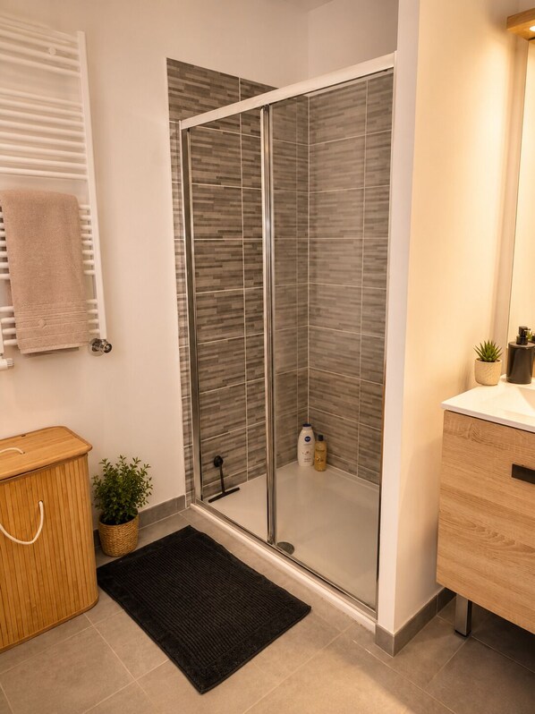 A modern bathroom with a glass shower enclosure, a wooden vanity, a black bath mat, and a potted plant.