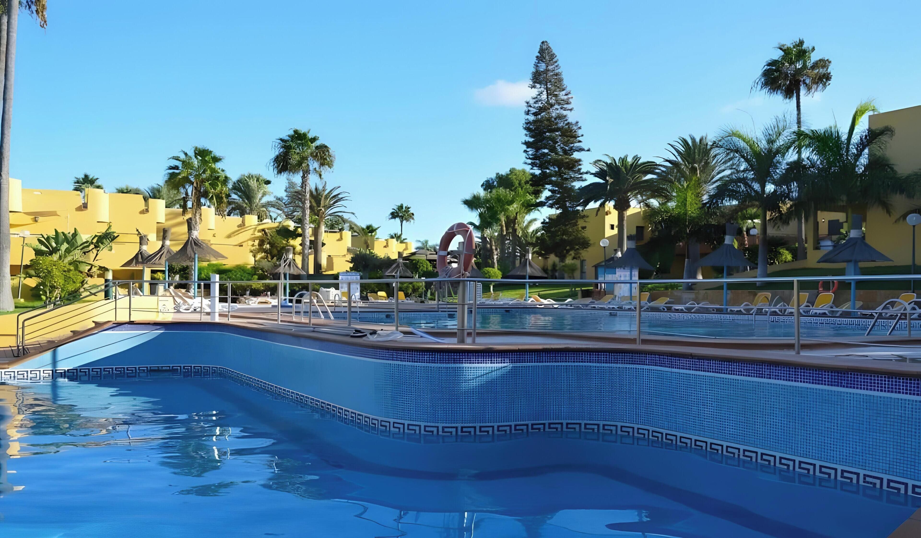 A swimming pool with a decorative edge, lounge chairs, palm trees, and yellow buildings in the background.