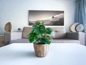 Una sala de estar con un sofá, una mesa de centro, una planta en maceta y una obra de arte en la pared que muestra a un surfista.
