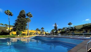 Una piscina bajo un cielo azul despejado, rodeada de palmeras y edificios amarillos.