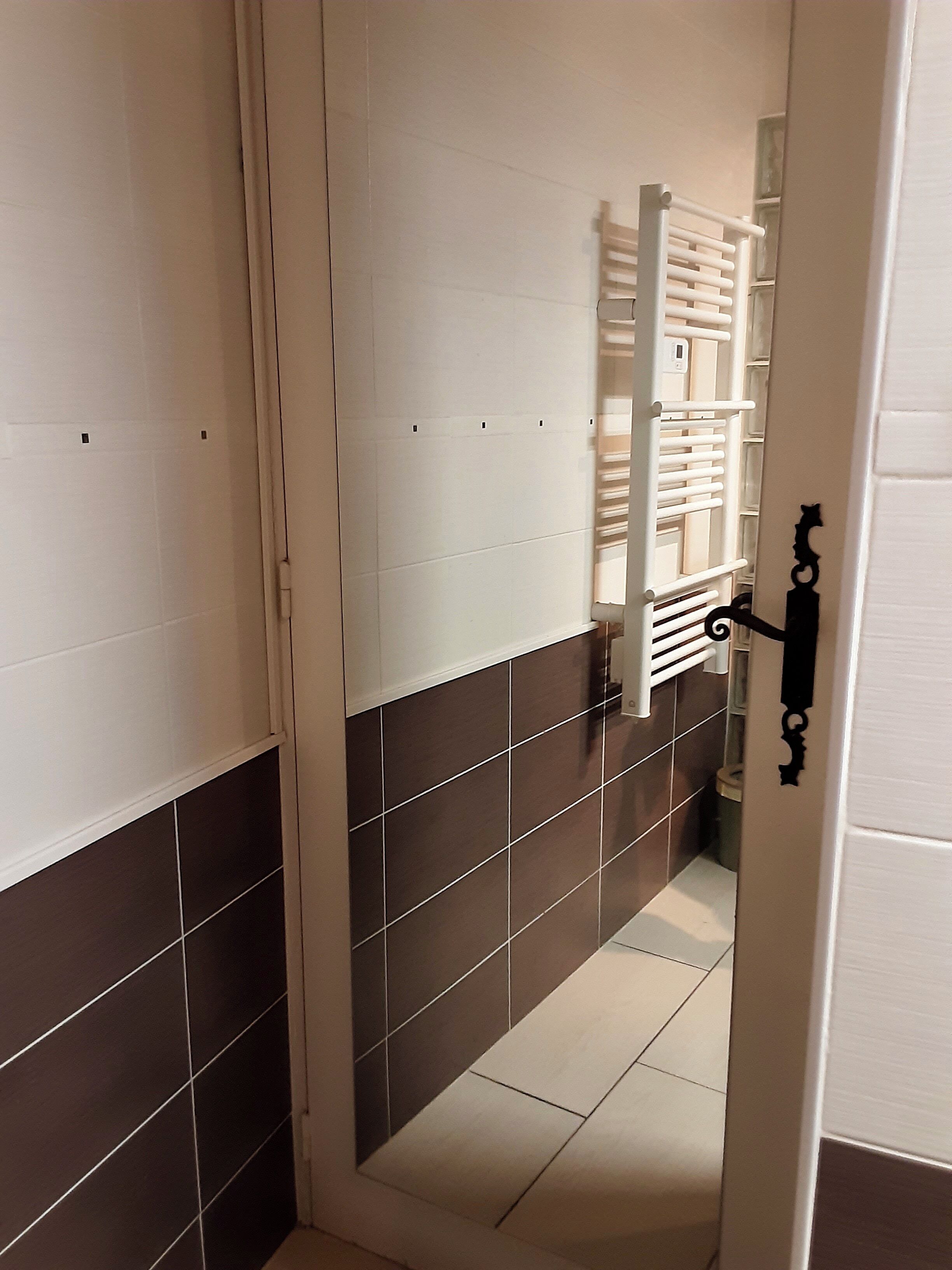 Een smalle gang met een betegelde vloer, een witte deur met een zwarte hendel en een witte radiator aan de muur.