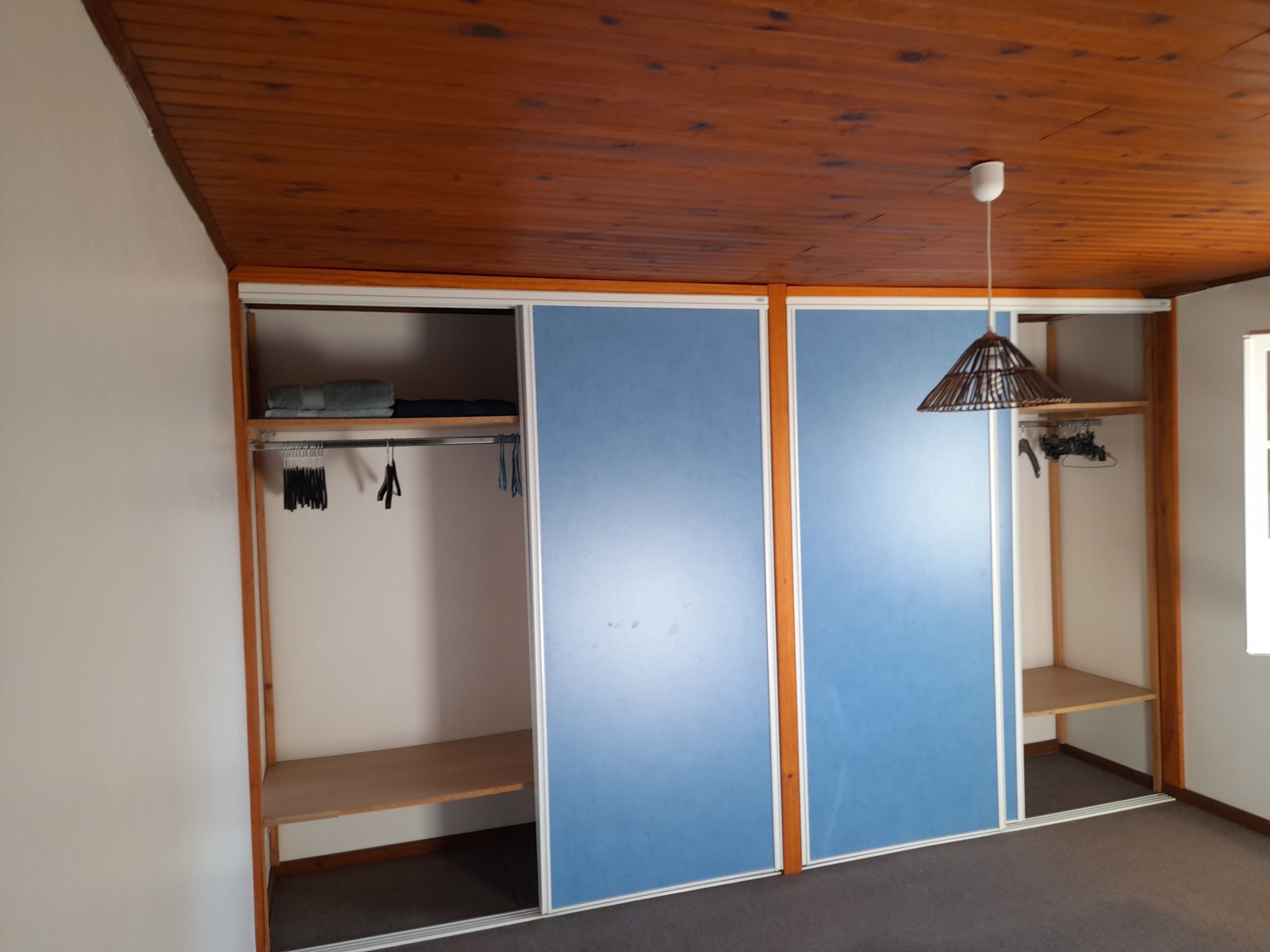 Una stanza con soffitto in legno, un armadio con porta blu, una lampada a sospensione e una finestra.
