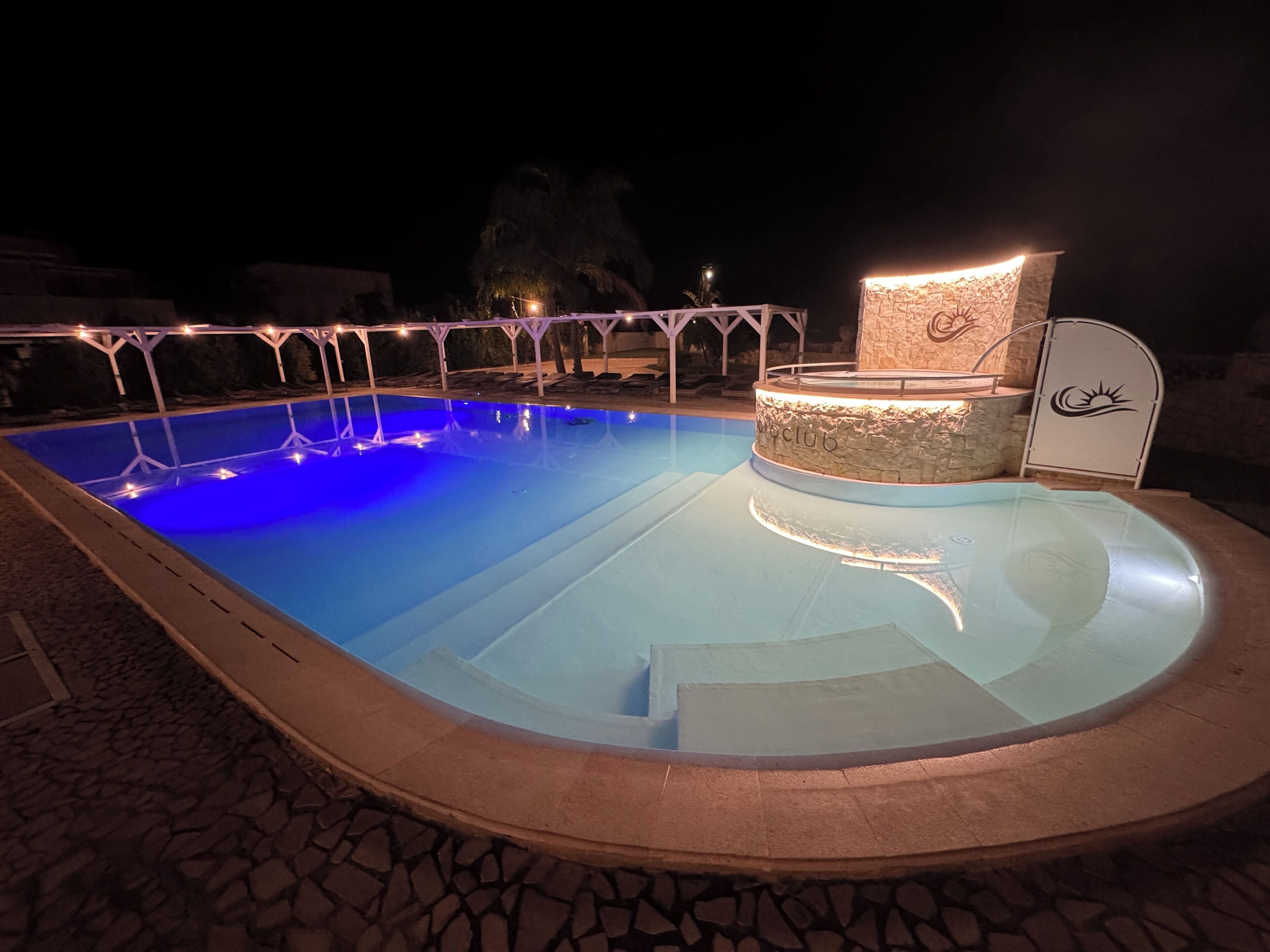 A swimming pool with a stone surround, a lounge area with a stone wall, and a covered seating area with a logo.