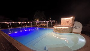Une piscine entourée de pierres, un espace détente avec un mur en pierre et un coin salon couvert orné d’un logo.