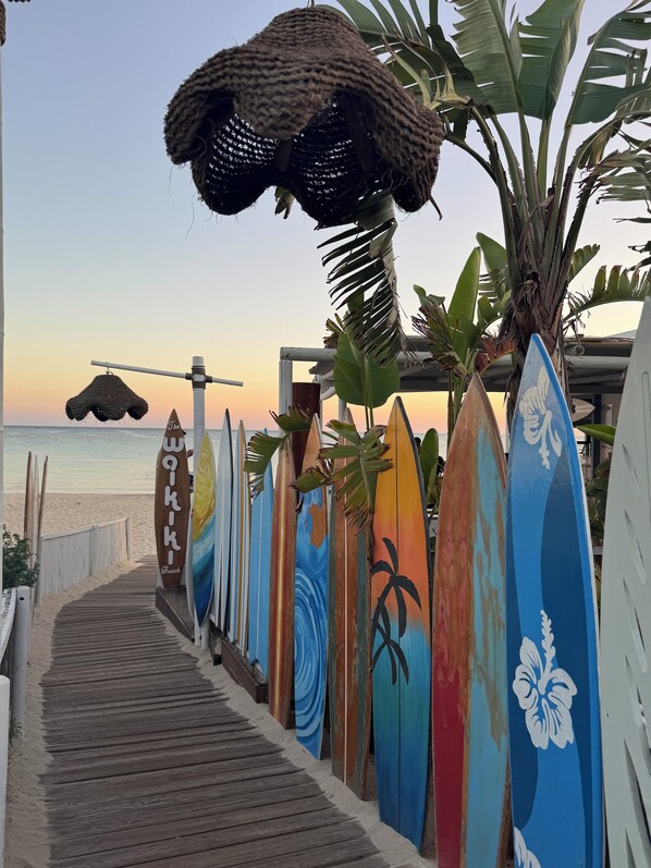 Ein hölzerner Steg führt zu einem Strand, an dem Surfbretter aufgereiht sind.