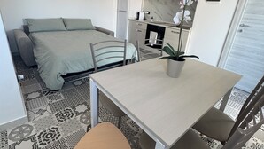 Un espace de vie compact comprenant un lit, une table à manger avec des chaises, un coin cuisine et une orchidée en pot.