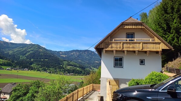 A house with a balcony, a car parked in front, and a mountainous landscape in the background.