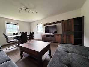 Una sala de estar moderna con una mesa de comedor de madera, sillas negras, un sofá gris, un televisor sobre un mueble de madera y una lámpara en el techo.