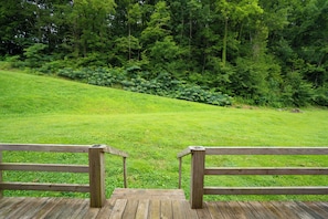 A wooden deck with a view of a grassy hill and dense forest. - 🍃 Quaint Cabin on Cosby Creek 🏞️ Creekside Farm, Hot Tub & Park Access (Cosby)