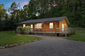 A wooden cabin with a deck, surrounded by a gravel driveway and a grassy area. - 🍃 Quaint Cabin on Cosby Creek 🏞️ Creekside Farm, Hot Tub & Park Access (Cosby)